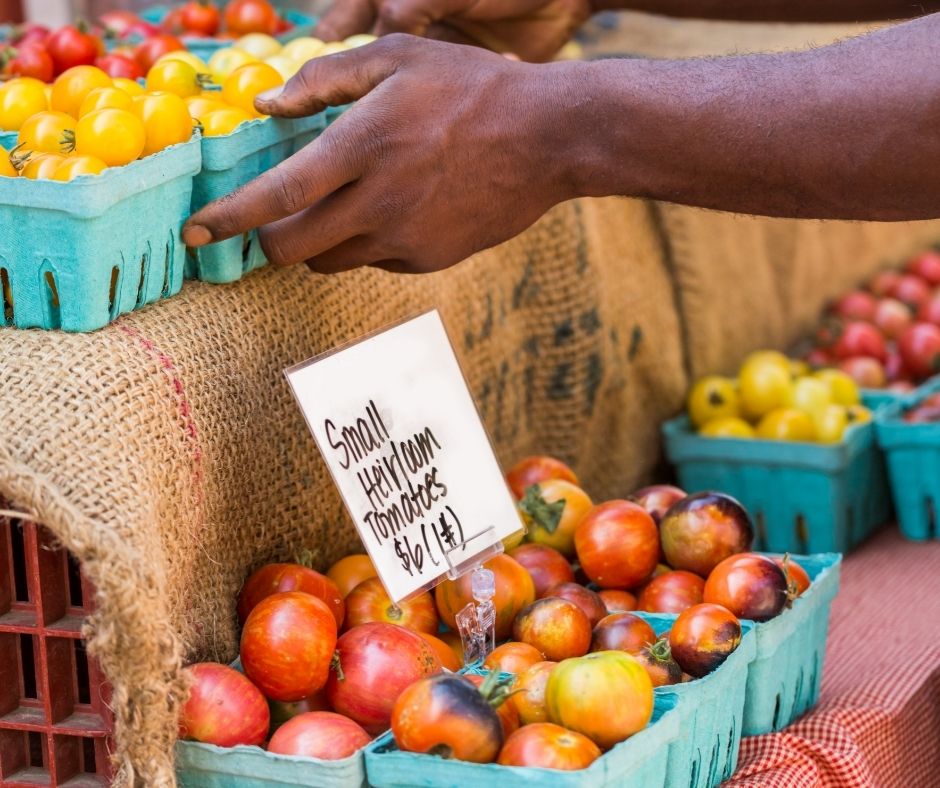 A farmers market stall featuring heirloom tomatoes, and a persons hands reaching in to pick up a container of them.
